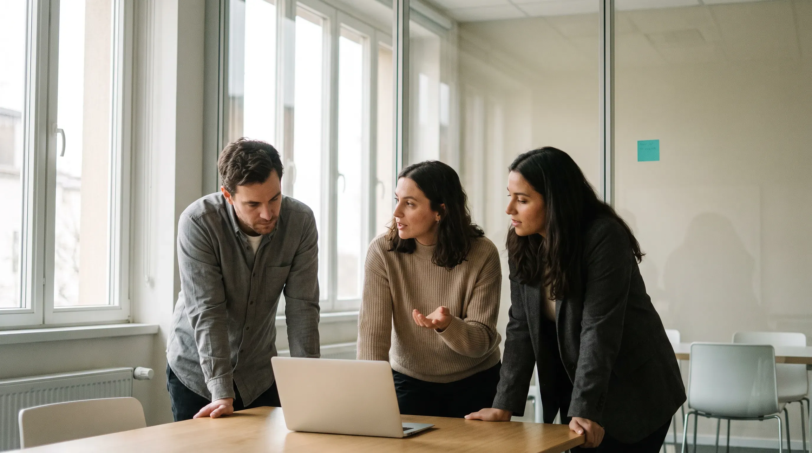 Three colleagues in a calm, light-filled office lean over a laptop mid-discussion, none looking at the camera, with a single small turquoise sticky note on the glass wall behind them.