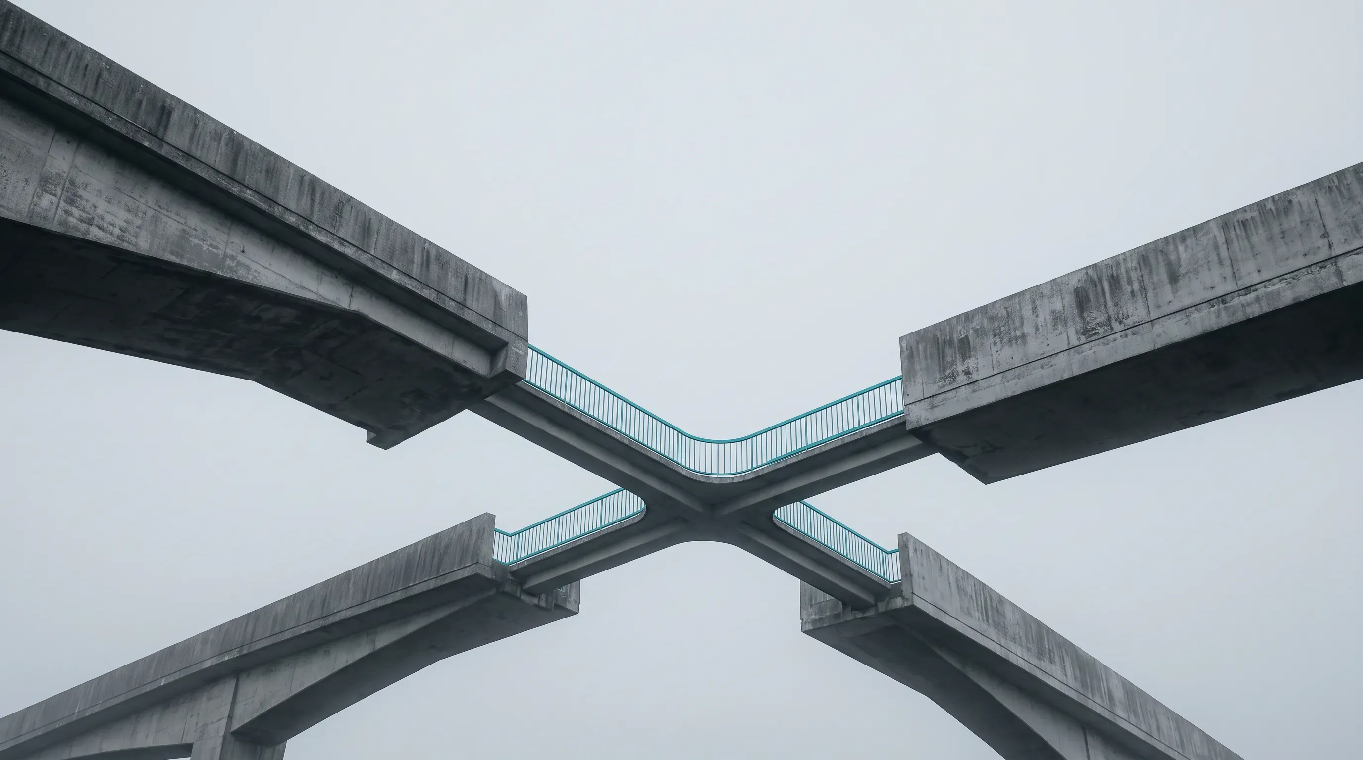 Four separate concrete bridge ends approach from different directions under a flat grey sky, joined only by one slender crossing walkway whose thin turquoise handrail is the single color in the frame.